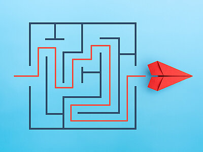 Ein Labyrinth mit einem roten Pfad, der den Weg durch das Labyrinth zeigt. Rechts neben dem Labyrinth liegt ein rotes Papierflugzeug. Das Labyrinth und das Flugzeug sind auf einem hellblauen Hintergrund platziert.