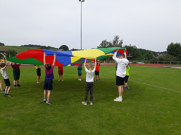 Unser Motto zum Sportfest: "Auf zu neuen Galaxien!" Kinder spielen mit einem großen, bunten Schwungtuch auf einer Rasenfläche. Sie heben es gemeinsam in die Luft.