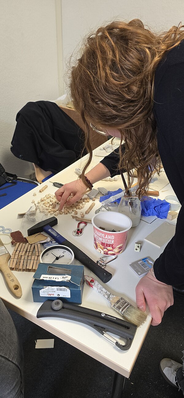 Eine Person mit lockigen Haaren beugt sich über einen Tisch voller Werkzeuge und Materialien und sortiert kleine Holzperlen für ein handwerkliches Projekt.