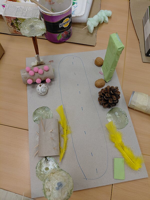 Auf einer Pappplatte ist eine vereinfachte Landschaft gestaltet: In der Mitte ist eine blaue Straße eingezeichnet, links und rechts stehen kleine Objekte aus Walnussschalen, Federn, Zapfen, Papprollen und glänzenden runden Formen, die wie Bäume und Häuser wirken.
