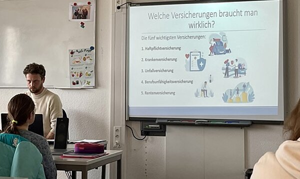 Welche Versicherungen braucht man wirklich? Herr Naun sitzt am Lehrerpult vor der Klasse, hinter ihm wird seine Präsentation an die Wand gebeamt.