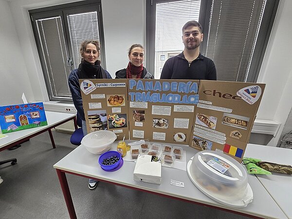Drei Azubis stehen hinter einem Tisch, auf dem eine Präsentation mit dem Titel 'PANADERIA TRIGUCIOLLO' aufgebaut ist. Die Präsentation besteht aus einem großen, aufgestellten Karton mit mehreren Fotos, Textfeldern und einer Flagge. Auf dem Tisch befinden sich verschiedene Lebensmittel, darunter ein Teller mit Oliven, mehrere kleine Behälter mit Backwaren, ein Glas mit gelber Flüssigkeit und ein abgedeckter Teller. Im Hintergrund sind zwei große Fenster und ein weiteres Präsentationsplakat auf einem anderen Tisch zu sehen.