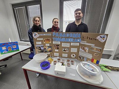 Drei Azubis stehen hinter einem Tisch, auf dem eine Präsentation mit dem Titel 'PANADERIA TRIGUCIOLLO' aufgebaut ist. Die Präsentation besteht aus einem großen, aufgestellten Karton mit mehreren Fotos, Textfeldern und einer Flagge. Auf dem Tisch befinden sich verschiedene Lebensmittel, darunter ein Teller mit Oliven, mehrere kleine Behälter mit Backwaren, ein Glas mit gelber Flüssigkeit und ein abgedeckter Teller. Im Hintergrund sind zwei große Fenster und ein weiteres Präsentationsplakat auf einem anderen Tisch zu sehen.