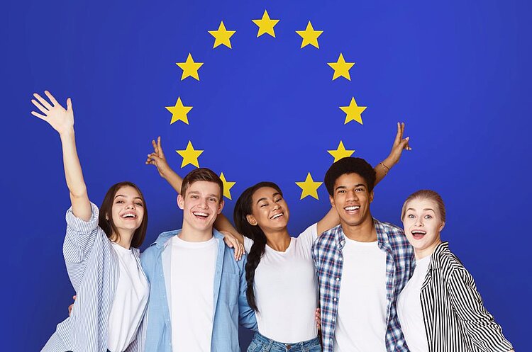 Unterwegs in Europa Eine fröhliche Gruppe junger Menschen unterschiedlicher Herkunft steht vor einer großen EU-Flagge mit gelben Sternen auf blauem Hintergrund. Sie lachen, umarmen sich und heben die Arme in die Luft, während sie in die Kamera blicken.