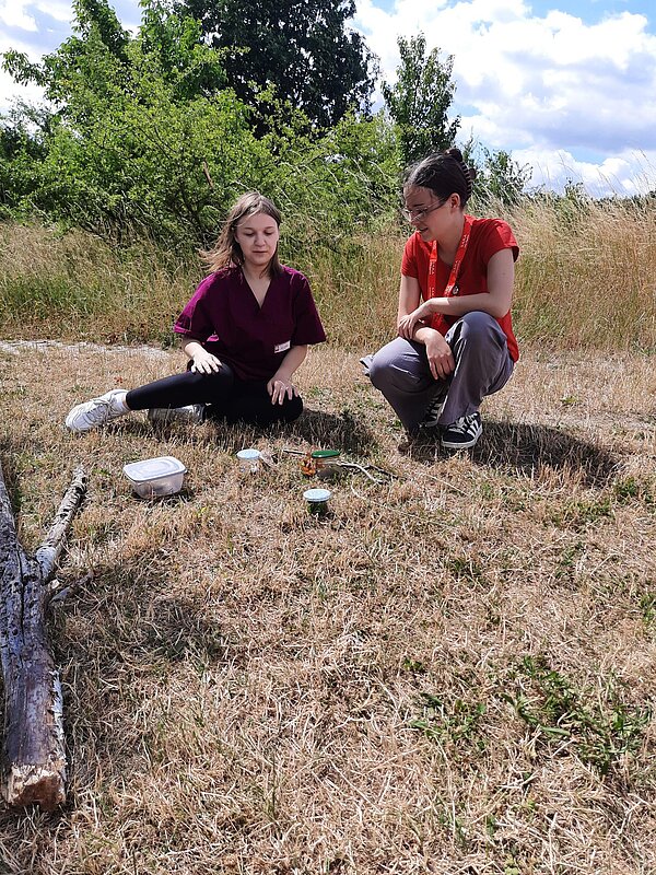 Ein Sinneskreis am Goitzsche-Strand: Lernen, Fühlen, Erleben! Zwei Frauen sitzen auf einer trockenen Wiese und beobachten kleine Dosen mit Naturmaterialien. Im Hintergrund Bäume und Gras.