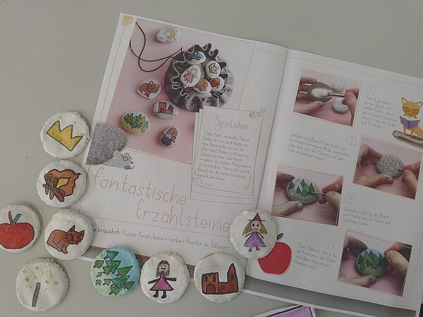 Damit der Herbst nicht zu traurig ist: Unsere Spielempfehlungen Ein Heft mit der Anleitung zum Basteln von Spielen liegt auf einem weißen Tisch, darum selbst bemalte Steine.