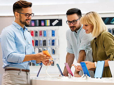Drei Personen stehen in einem Elektronikgeschäft und betrachten verschiedene Smartphones. Der Mann links, der ein hellblaues Hemd und eine Brille trägt, hält ein orangefarbenes Smartphone in der Hand und erklärt etwas. Das Paar rechts, ein Mann mit Brille und hellblauem Hemd sowie eine Frau mit blonden Haaren und grünem Oberteil, schauen interessiert auf die ausgestellten Smartphones in verschiedenen Farben wie Blau, Rosa und Schwarz. Im Hintergrund sind Regale mit weiteren Smartphone-Verpackungen zu sehen. Alle drei wirken freundlich und engagiert.