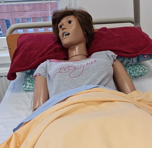 Pflege trifft Soziales Eine realistisch aussehende Pflegepuppe liegt in einem Krankenhausbett. Sie trägt ein graues T-Shirt mit rosa Schriftzug „Loves you“ und ist mit einer gelben Decke zugedeckt. Der Kopf der Puppe ist leicht zur Seite geneigt, der Mund geöffnet. Im Hintergrund ist ein Fenster mit Blick auf ein rotes Gebäude zu sehen.