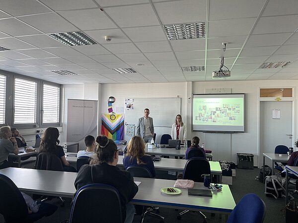 Career Opportunities an der Euro Akademie - Einblicke in die Berufswelt Zwei Referierende stehen vor einer Präsentation mit Bildern von Mitarbeitenden. Im Raum sitzen Schülerinnen und Schüler, links ist ein Roll-up mit dem Logo von Hengeler Mueller zu sehen. An der Wand hängt eine große Progress-Pride-Flagge.