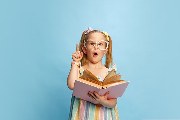 Ein kleines Mädchen in einem bunten Sommerkleid steht vor einem hellblauen Hintergrund. Es trägt eine zu große Brille und hat zwei Zöpfe. Das Mädchen hält ein Buch in der Hand und erhebt ihren rechten Zeigefinger, während sie den Mund zu einem "O" formt.