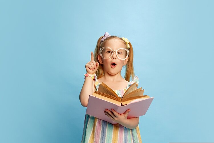 Ein kleines Mädchen in einem bunten Sommerkleid steht vor einem hellblauen Hintergrund. Es trägt eine zu große Brille und hat zwei Zöpfe. Das Mädchen hält ein Buch in der Hand und erhebt ihren rechten Zeigefinger, während sie den Mund zu einem "O" formt.