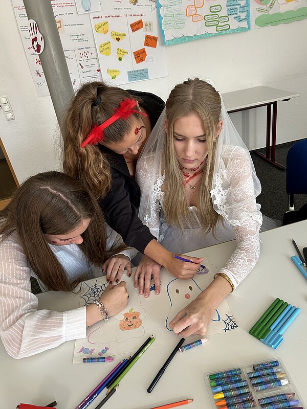 Nahaufnahme von drei Lernenden, die gemeinsam Halloween-Motive wie Geist, Kürbis und Spinnennetz kolorieren.