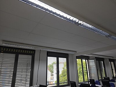 Deckenleuchte mit langem, rechteckigem Lichtband in einem Büroraum. Die Fensterfront zeigt Lamellenjalousien, durch die Tageslicht und grüne Bäume sichtbar sind. Mehrere Computerarbeitsplätze stehen unter der Leuchte.