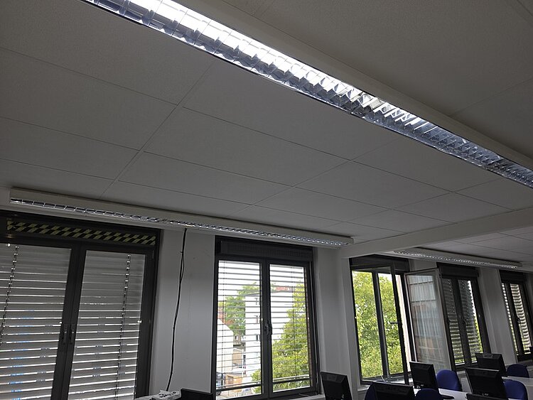 Deckenleuchte mit langem, rechteckigem Lichtband in einem Büroraum. Die Fensterfront zeigt Lamellenjalousien, durch die Tageslicht und grüne Bäume sichtbar sind. Mehrere Computerarbeitsplätze stehen unter der Leuchte.