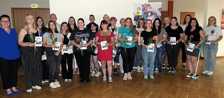 Zu sehen ist eine große Gruppe von Schülerinnen und Schülern, die eine Blume und einen Flyer in der Han halten. Die Gruppe steht in einem Raum mit Parkettboden.