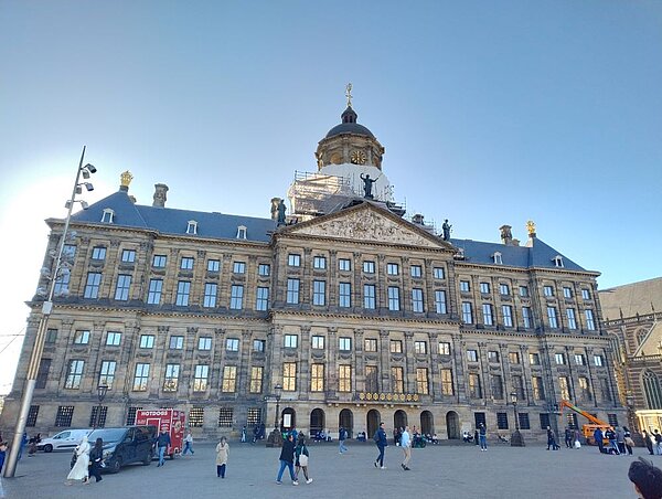 Das historische Royal Palace (Königspalast) in Amsterdam mit klassizistischer Fassade und einem großen Platz davor. Passanten gehen vorbei, und die Sonne scheint auf das Gebäude.
