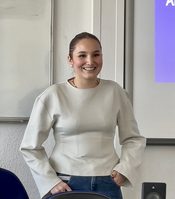 Eine junge Frau steht lächelnd vorne im Klassenraum neben einer Präsentationsfläche.
