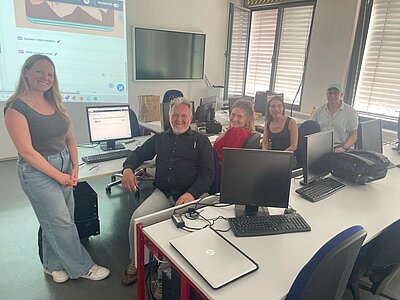 Eine Gruppe von vier Erwachsenen sitzt in einem Computerraum an Tischen mit Monitoren und Laptops. Eine Frau steht links daneben und lächelt in die Kamera. Im Hintergrund sind Fenster mit Jalousien sowie eine große Leinwand mit einer Präsentation zu sehen.