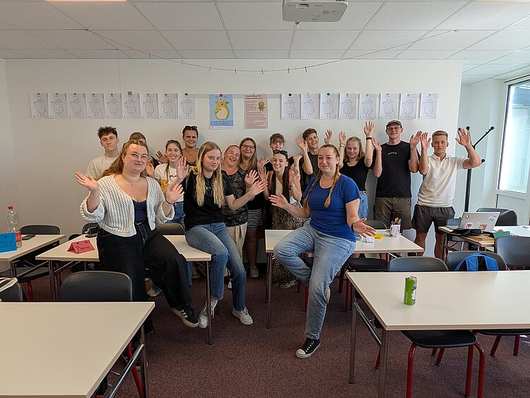 Teambuilding mit Herz Eine Gruppe von etwa 17 Personen sitzt und steht lachend in einem Klassenraum und hebt gemeinsam die Hände.