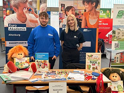 Zwei Personen stehen an einem Infostand der Euro Akademie Altenburg mit Roll-ups zu den Ausbildungen Kinderpflegerin und Erzieherin; auf dem Tisch liegen Infomaterialien, Stifte, Süßigkeiten, ein Buch zu Gefühlen und eine Stoffpuppe.