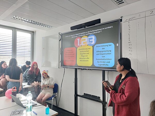 Eine junge Frau hält ein Tablet und steht vor einer digitalen Tafel, auf der verschiedene Themenbereiche wie Pflege, Versorgung und Betreuung von Menschen angezeigt werden. Im Hintergrund sitzen und stehen weitere Personen und verfolgen aufmerksam die Präsentation.