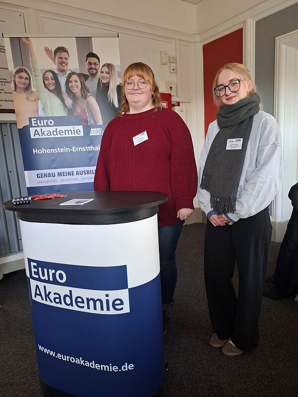 Tag der offenen Tür an der Euro Akademie Hohenstein-Ernstthal: Zwei Personen stehen vor einem Werbeständer der Euro Akademie Hohenstein-Ernstthal. Beide tragen Namensschilder und lächeln in die Kamera. Der Ständer ist blau und zeigt das Logo sowie Fotos von Menschen.