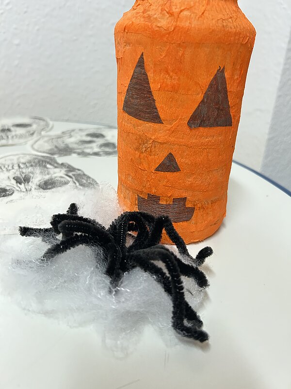 Selbstgebastelte Halloween-Deko: orange bemalte Flasche mit Kürbisgesicht, daneben eine schwarze Pfeifenreiniger-Spinne auf Kunstspinnennetz.