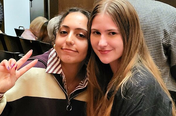 Zwei Schülerinnen der Euro Akademie Hannover sitzen im Opernhaus Hannover während ihres Besuchs des Balletts „Goldberg“. Die Jugendlichen lächeln in die Kamera; eine zeigt ein Peace-Zeichen. Beide sitzen eng beieinander in einer Zuschauerreihe. Hinter ihnen sind weitere Besucherinnen und Besucher des Opernhauses zu erkennen. Die warme Beleuchtung und die elegante Atmosphäre des Saals vermitteln eine konzentrierte, kulturelle Stimmung.