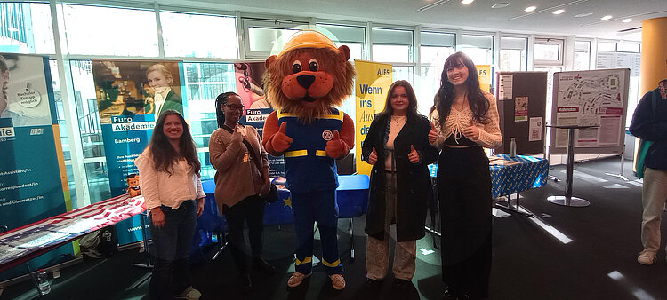 Vor dem Messestand der Euro Akademie stehen lächelnd vier junge Frauen, in ihrer Mitte ein Mensch in Löwenkostüm mit einer Weste des Technischen Hilfswerks.