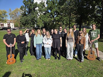 Gruppe von 16 Personen steht lächelnd im Grünen vor Bäumen und einem Gebäude, zwei Personen halten Gitarren; blauer Himmel, entspannte Stimmung.