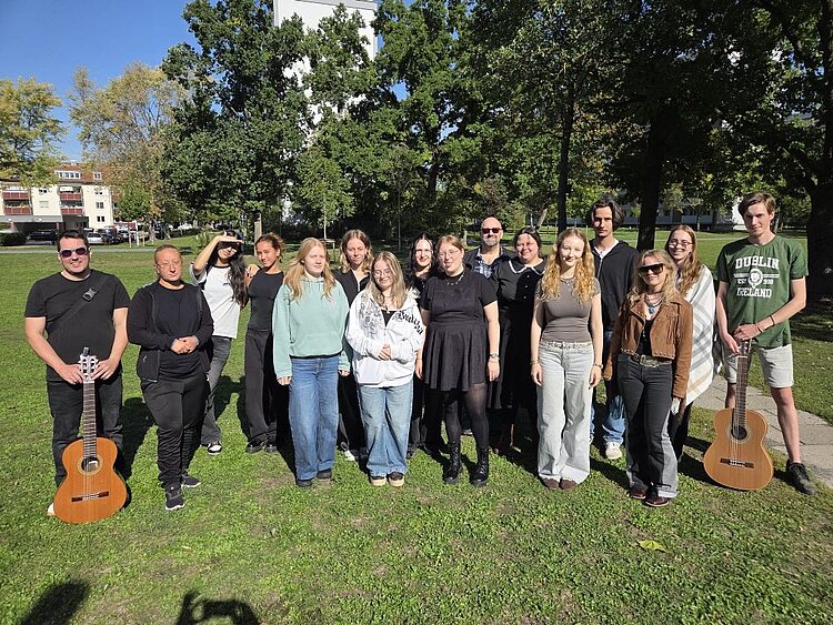 Gruppe von 16 Personen steht lächelnd im Grünen vor Bäumen und einem Gebäude, zwei Personen halten Gitarren; blauer Himmel, entspannte Stimmung.