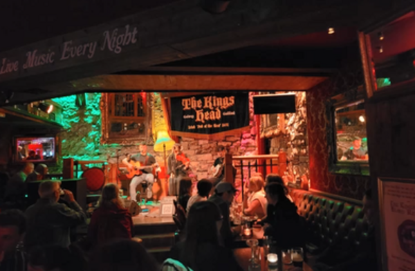 Ein irischer Pub der Stadt Galway von innen mit einer Bar, vielen Tischen und Stühlen und Leuten, die etwas trinken. Dabei ist es relativ dunkel.
