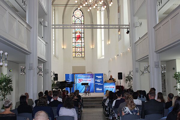 Euro Akademie Görlitz und Zittau: Feierliche Exmatrikulation Vor dem Altar in einer Kirche ist ein Podium aufgebaut und verschiedene mobile Wände mit dem Euro Akademie Logo. Am Podium steht eine Frau und hält eine Rede.