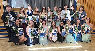 Vor einer Bühne steht und kniet eine Gruppe junger Menschen in festlichen Kleidern. Sie halten Blumen und Zeugnismappen in den Händen.