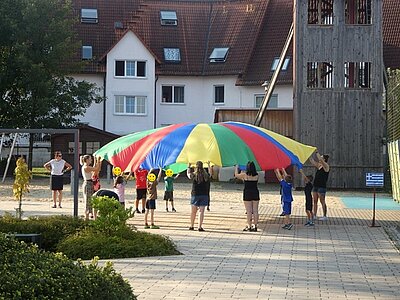 Kinder und Auszubildende stehen draußen auf dem Hof und spielen mit dem großen, bunten Schwingtuch.
