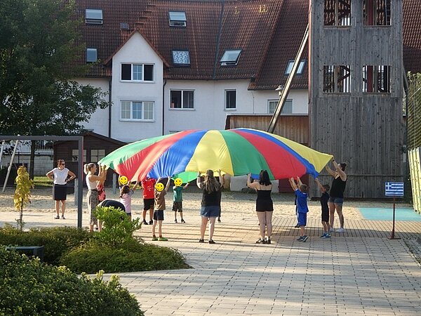 Kinder und Auszubildende stehen draußen auf dem Hof und spielen mit dem großen, bunten Schwingtuch.