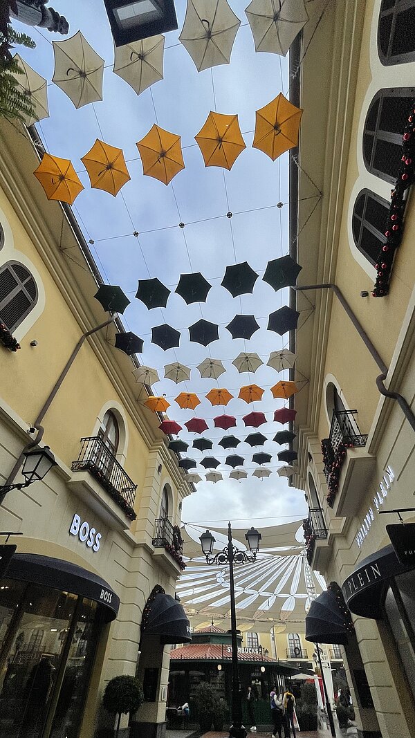 Offenes Einkaufszentrum mit hellen Fassaden und Balkonen, darüber ein Dach aus vielen aufgehängten Schirmen in Gelb, Weiß, Rot und Dunkelgrün, im unteren Bildrand Ladenfronten wie „BOSS“.