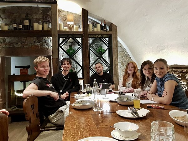 Alumnitreffen 2025 Drei männliche und drei weibliche ehemalige Auszubildende der Euro Akademie Aschaffenburg sitzen an einem Tisch im Restaurant und posieren lächelnd für ein Gruppenfoto. Vor ihnen stehen leere Teller, Besteck, Gläser mit verschiedenen Getränken und eine kleine Tischlampe.