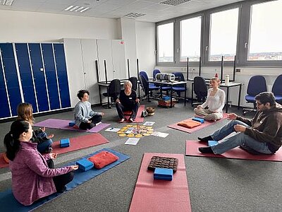 Eine Gruppe von sechs Personen sitzt auf Yogamatten in einem Halbkreis in einem modernen Raum mit großen Fenstern. Eine ältere Frau in der Mitte leitet die Meditation, während die Teilnehmenden mit geschlossenen Augen oder entspannten Gesichtsausdrücken sitzen. In der Mitte des Kreises liegen Karten und kleine Gegenstände