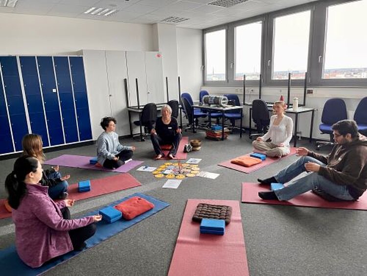 Eine Gruppe von sechs Personen sitzt auf Yogamatten in einem Halbkreis in einem modernen Raum mit großen Fenstern. Eine ältere Frau in der Mitte leitet die Meditation, während die Teilnehmenden mit geschlossenen Augen oder entspannten Gesichtsausdrücken sitzen. In der Mitte des Kreises liegen Karten und kleine Gegenstände