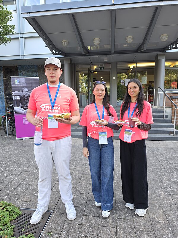 Vor einem Gebäude stehen drei Personen in pinken T-Shirts mit Ausweisbändern um den Hals. Die Personen halten Sandwiches und Getränke in den Händen.