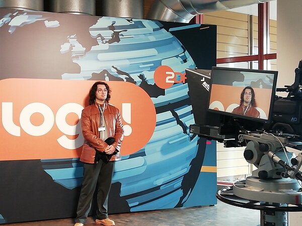 Ein junger Mann steht vor einer Wand mit der Aufschrift logo ZDF und wird dabei gefilmt.