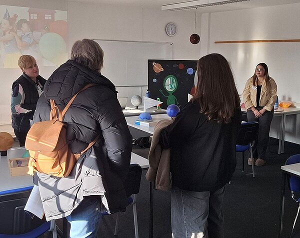 Tag der offenen Tür an der Euro Akademie Hohenstein-Ernstthal: Eine Gruppe von Erwachsenen steht und sitzt in einem Klassenzimmer. Im Hintergrund sind eine Tafel und ein Plakat mit einem Weltraum-Motiv zu sehen. Zwei Personen tragen Jacken und Rucksäcke, während eine weitere Person vorne an einem Tisch steht und spricht.