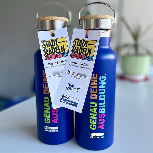 Zwei Thermoskannen in blau, mit buntem Schriftzug "Genau Deine Ausbildung" und Logo der Euro Akademie. An beiden Flaschen hängen Zettel mit der Auszeichnung für Platz 1 und die beste Radlerin.