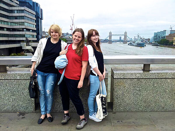 Drei junge lächelnde Frauen mit Taschen stehen auf einer Brücke, die über einen breiten Fluss führt. Es scheint die Themse zu sein, denn im Hintergrund ist die Tower Bridge zu sehen.