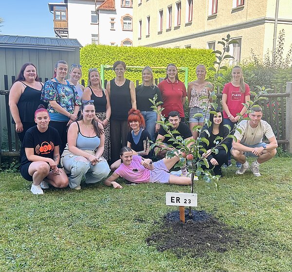 Grüner Schulstart 2025/26 an der Euro Akademie Altenburg Eine Gruppe von Menschen steht im Freien um einen kleinen Baum. Sie posieren lächelnd für ein Gruppenfoto. Der Baum hat ein Schild mit der Aufschrift "ER 23". Die Gruppe besteht aus jungen Erwachsenen und sie stehen auf einem Rasen mit einem Gebäude im Hintergrund.