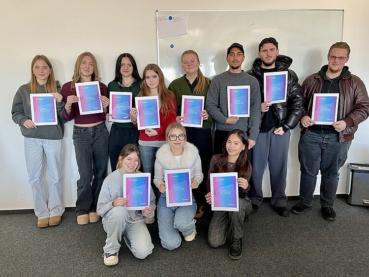 Die Gruppe von elf Teilnehmenden der Euro Akademie Aschaffenburg stehen und knien teilweise vor einer weißen Wand mit einem Whiteboard. Alle halten jeweils eine Urkunde mit einem Farbverlauf von Blau zu Pink, die den Europass "Mobilität" des Erasmus+-Programms symbolisiert in den Händen. Die Personen tragen unterschiedliche, legere, moderne Kleidung, darunter Pullover, Jacken und Jeans. Die Szene zeigt die feierliche Übergabe der Europässe an die Teilnehmenden, die sie freudig entgegen nehmen.