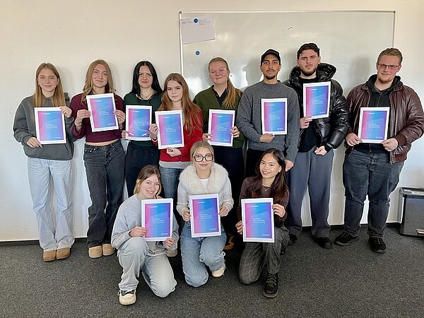 Die Gruppe von elf Teilnehmenden der Euro Akademie Aschaffenburg stehen und knien teilweise vor einer weißen Wand mit einem Whiteboard. Alle halten jeweils eine Urkunde mit einem Farbverlauf von Blau zu Pink, die den Europass "Mobilität" des Erasmus+-Programms symbolisiert in den Händen. Die Personen tragen unterschiedliche, legere, moderne Kleidung, darunter Pullover, Jacken und Jeans. Die Szene zeigt die feierliche Übergabe der Europässe an die Teilnehmenden, die sie freudig entgegen nehmen.