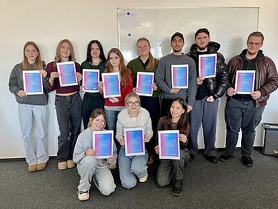Die Gruppe von elf Teilnehmenden der Euro Akademie Aschaffenburg stehen und knien teilweise vor einer weißen Wand mit einem Whiteboard. Alle halten jeweils eine Urkunde mit einem Farbverlauf von Blau zu Pink, die den Europass "Mobilität" des Erasmus+-Programms symbolisiert in den Händen. Die Personen tragen unterschiedliche, legere, moderne Kleidung, darunter Pullover, Jacken und Jeans. Die Szene zeigt die feierliche Übergabe der Europässe an die Teilnehmenden, die sie freudig entgegen nehmen.