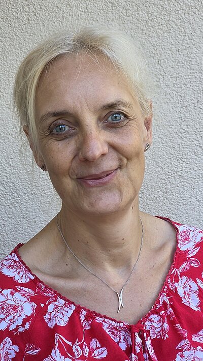 Porträtfoto der Mitarbeiterin Ilka Hofmann, die in einer rot-weißen Bluse, mit einer goldenen Kette um den hals freundlich in die Kamera lächelt.
